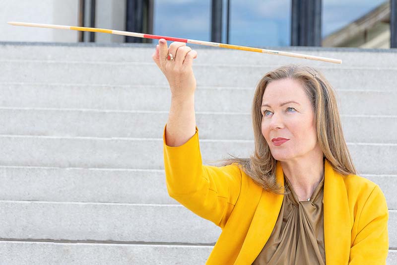 Porträt Frau auf einer Treppe mit gelbem Mantel und balanciert mit ein dünnes Stäbchen in der rechten Hand.